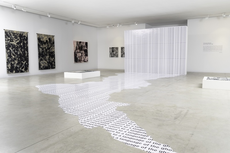  An installation shot in a white wall gallery. Five paintings hang on the far wall. In the foreground there is a text piece displayed on the gallery floor. 