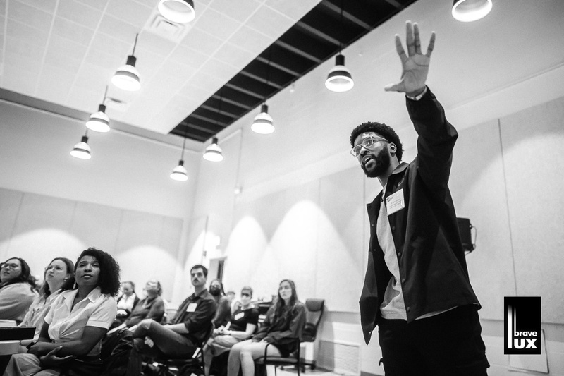  A black and white photo of a black male with short curly hair, a beard and glasses talking to an audience in a room. They are gesturing to something off camera as individuals look towards that spot with curiosity. 