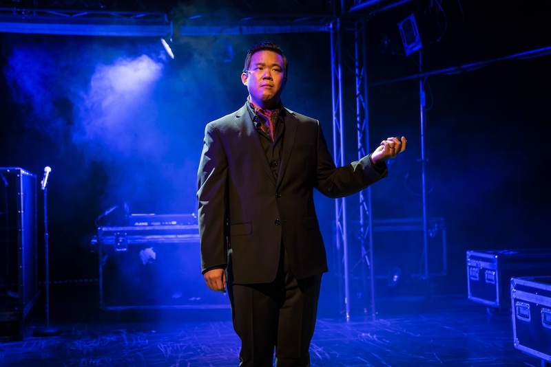  A still of a Filipino-Chinese cis male with short dark hair and olive skin standing on a blue stage. He is wearing a black suit. 