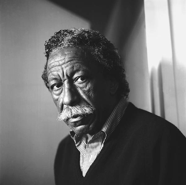  A black and white photo of an older Black man. He has a mustache and is wearing a checkered collared shirt. 