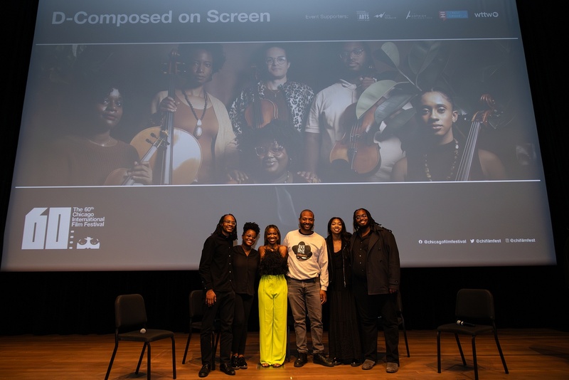  Six people stand in front of a projection screen that reads D-Composed on Screen. There are black chairs behind them and two microphones on the chairs. 
