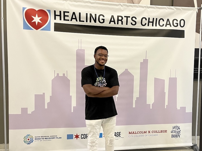  A Black Masc Person with short black locs and brown eyes stands in front of a banner that reads Healing Arts Collective. The banner has the outline of Chicago sketched on it. 