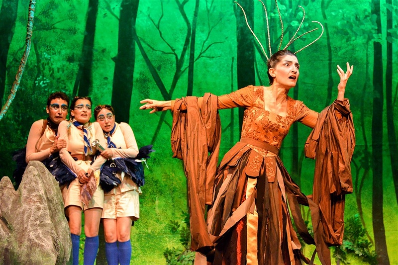  A performer strikes a pose while performing in an orange fringed dress. They have a wired crown on their head. Three other performers pose behind them. They are in a forest. 