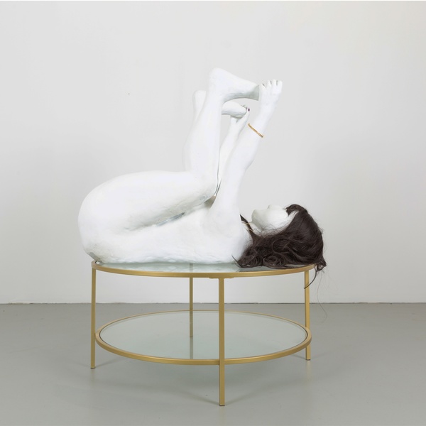  Photo of a sculpture in a white wall gallery space. The sculpture is of a human with no features, it has white skin and dark hair. It is laying with its back on a gold table with a glass top. The hands are holding the feet. 