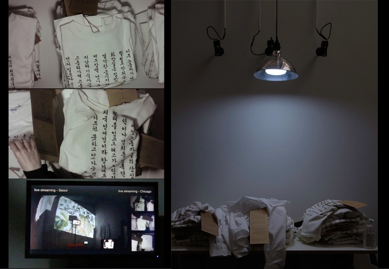  In a dark room lit by a suspended lamp, three piles of t-shirts with Korean calligraphy on display. On the wall three small camera face those t-shirts. 