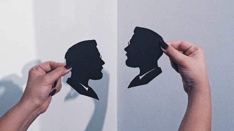  Two hands hold out two black portraits that are cut out by the artist. 
