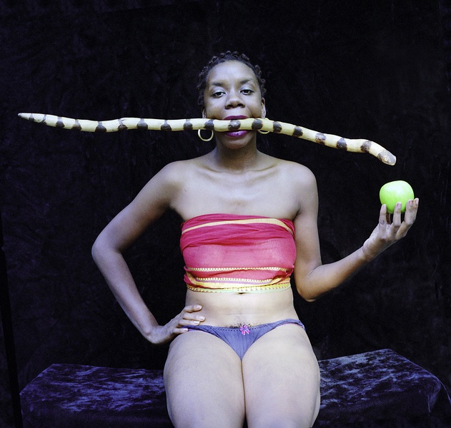  A portrait of a Black woman sitting on a blue velvet fabric. She is holding a green apple and has a yellow snake in her mouth. 