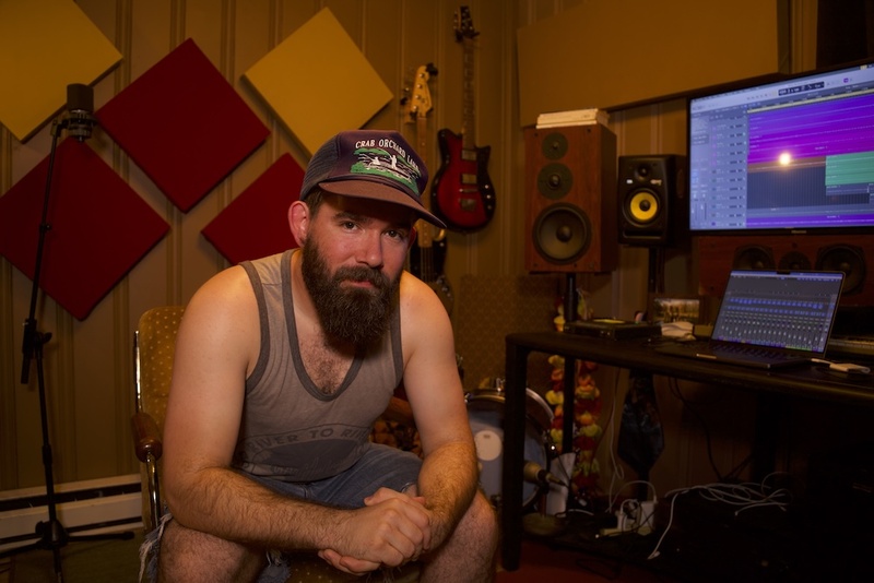  A medium built white man with short, light-brown hair and a dark brown, medium length, full beard poses and leans forward in a music studio. 