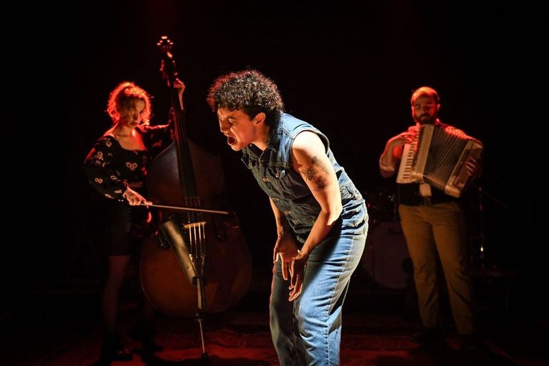  White, Queer, Non-binary Person with curly hair singing in a jean jacket and jean pants. A person playing the cello is on their left and a person with an accordion playing to their right. 