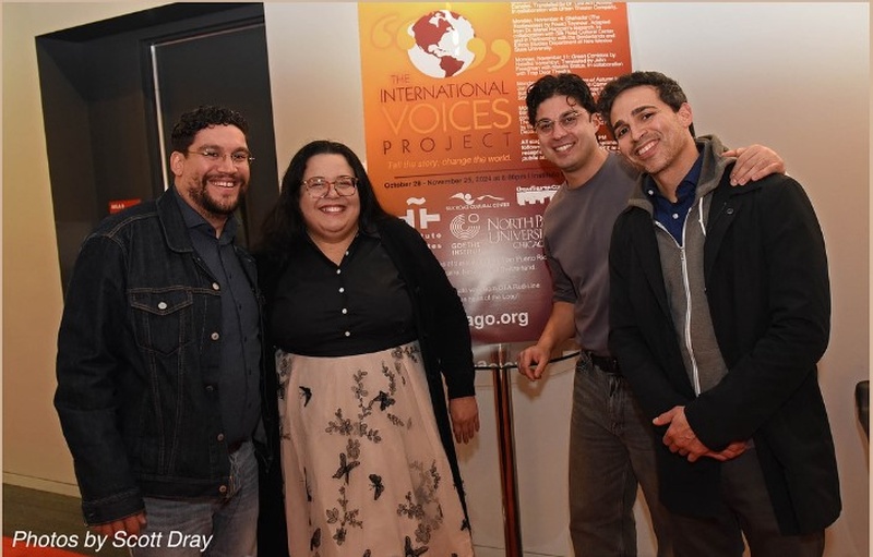  Four people stand, smiling at the camera in front of a poster that reads the international voices project. 