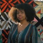 young woman with curly hair and dark brown skin stands in front of a colorful tapestry