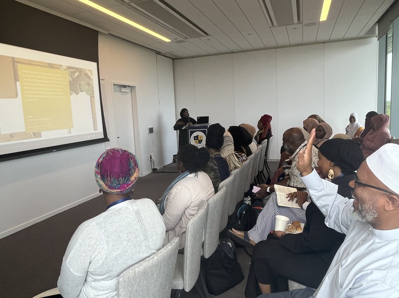  A Black Woman in a Hijab stands at a podium in a classroom in front of a slideshow presentation. Members of the audience have their hands raised. 