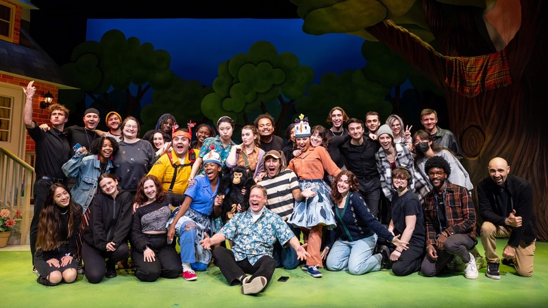 A cast photo of a handful of people who worked on a theater production. They are posed, smiling for a camera on green astroturf with faux trees and a porch in the background. 