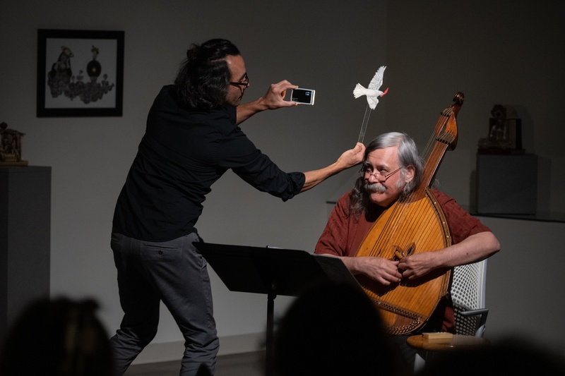  a biracial person with long black hair operates a small puppet of a white bird, flying it over the head of a musician with grey hair holding a wooden string instrument. 