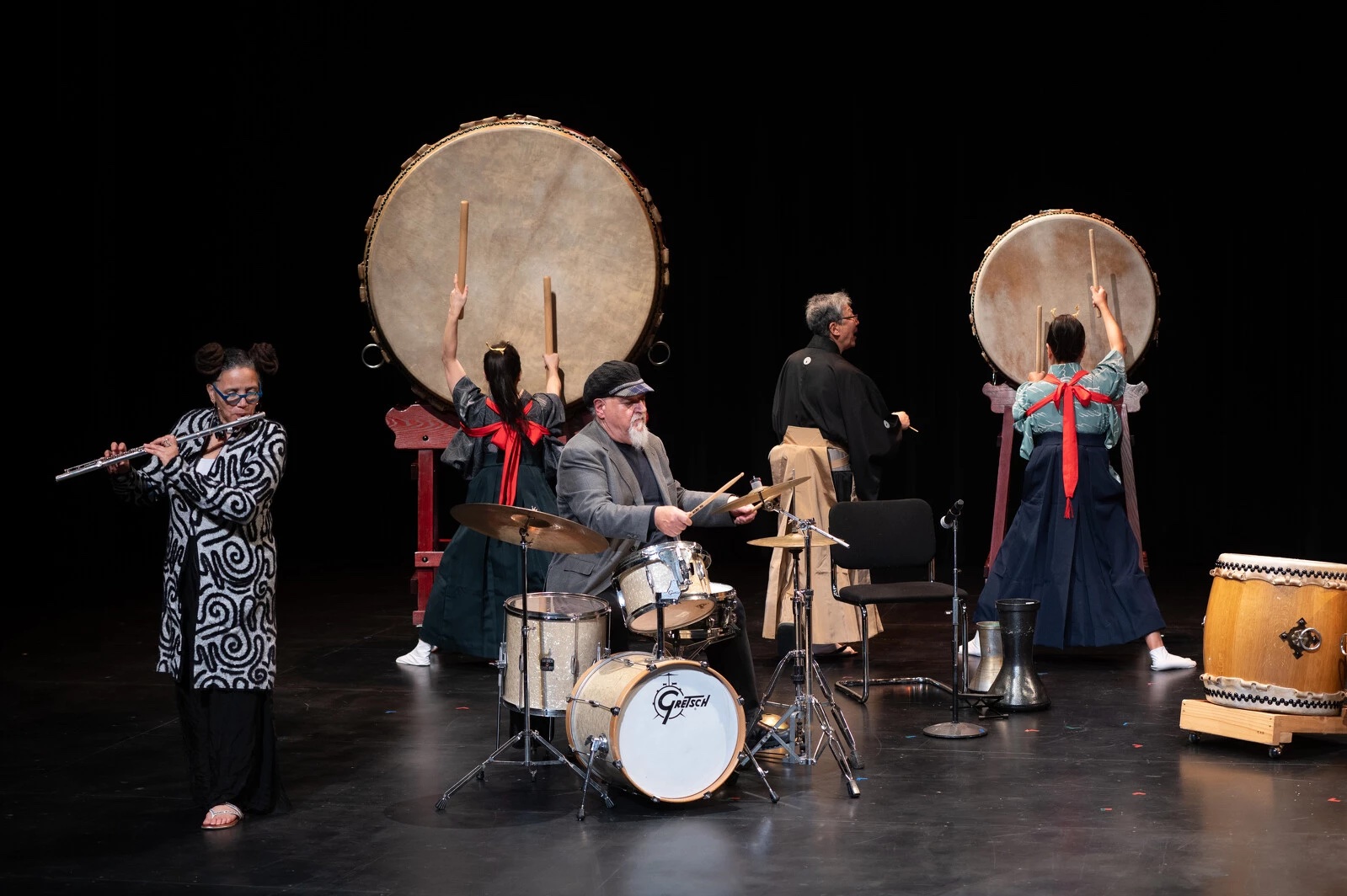 Image of performance group Reduction 11. The photo includes a drummer at center, a flutist on the left, two drummers in the background, and another musician facing the back wall.
